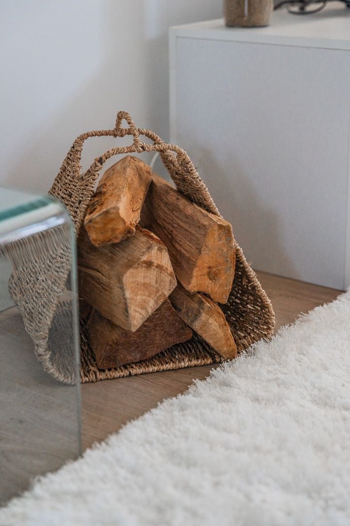 A natural woven basket holds neatly stacked pieces of firewood. The basket is positioned next to a glass-topped table, contrasting with a soft, light-colored rug that covers the floor.