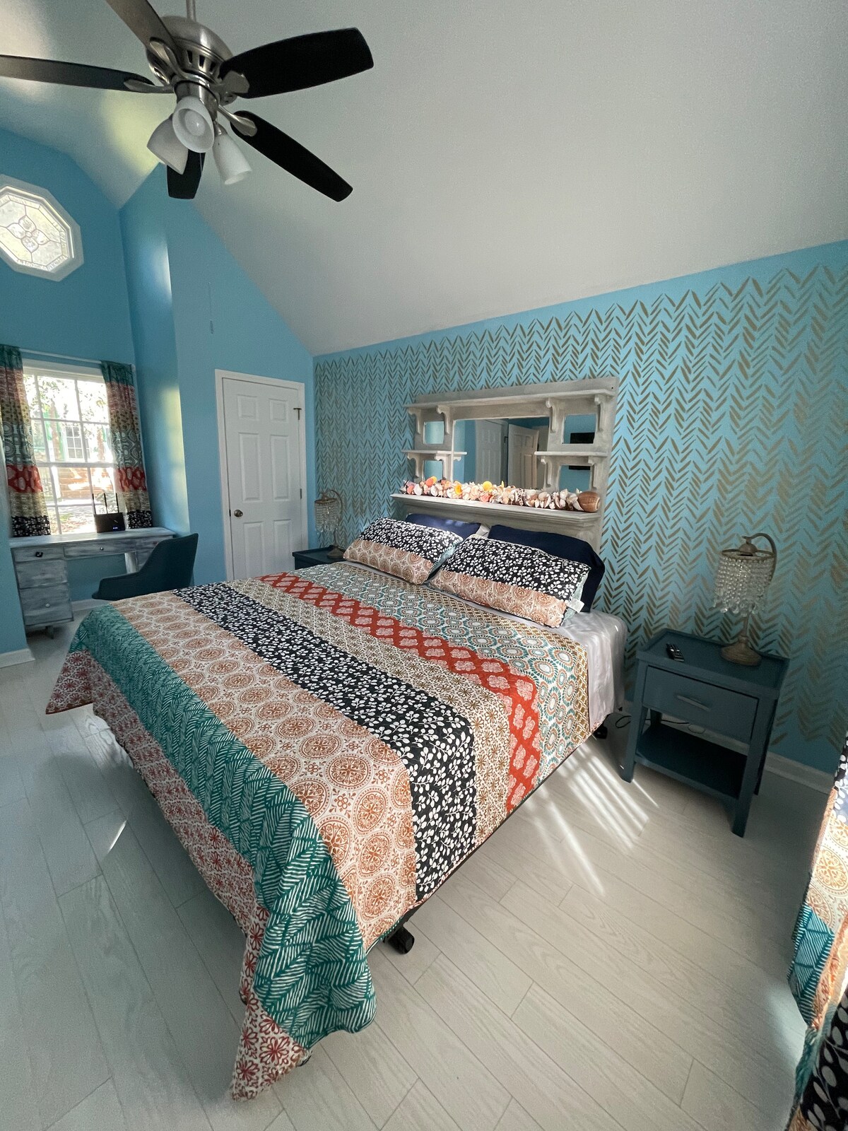 A bright bedroom features a spacious bed adorned with a colorful quilt, complemented by patterned throw pillows. The walls are painted in a soothing blue shade, and natural light enters through a window dressed with patterned curtains. A dresser with a mirror and a desk area are also visible.