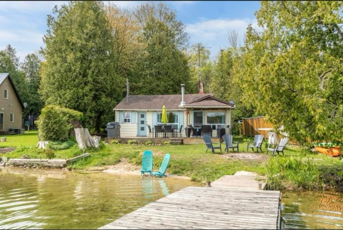 A rustic cabin is situated by the waters of William's Lake, surrounded by trees. The outdoor area features a wooden dock leading to the water, with two turquoise lounge chairs placed nearby. The cabin's porch is furnished with a dining table and a yellow umbrella.