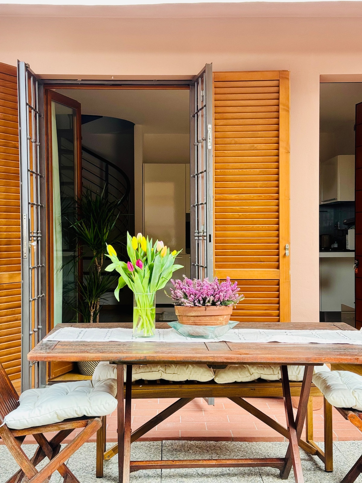 A wooden table is centered outside, adorned with a bouquet of flowers in a pot. Two chairs with soft cushions are positioned around the table. The background reveals an open doorway with slatted wood shutters and a glimpse of indoor greenery, enhancing the inviting atmosphere.