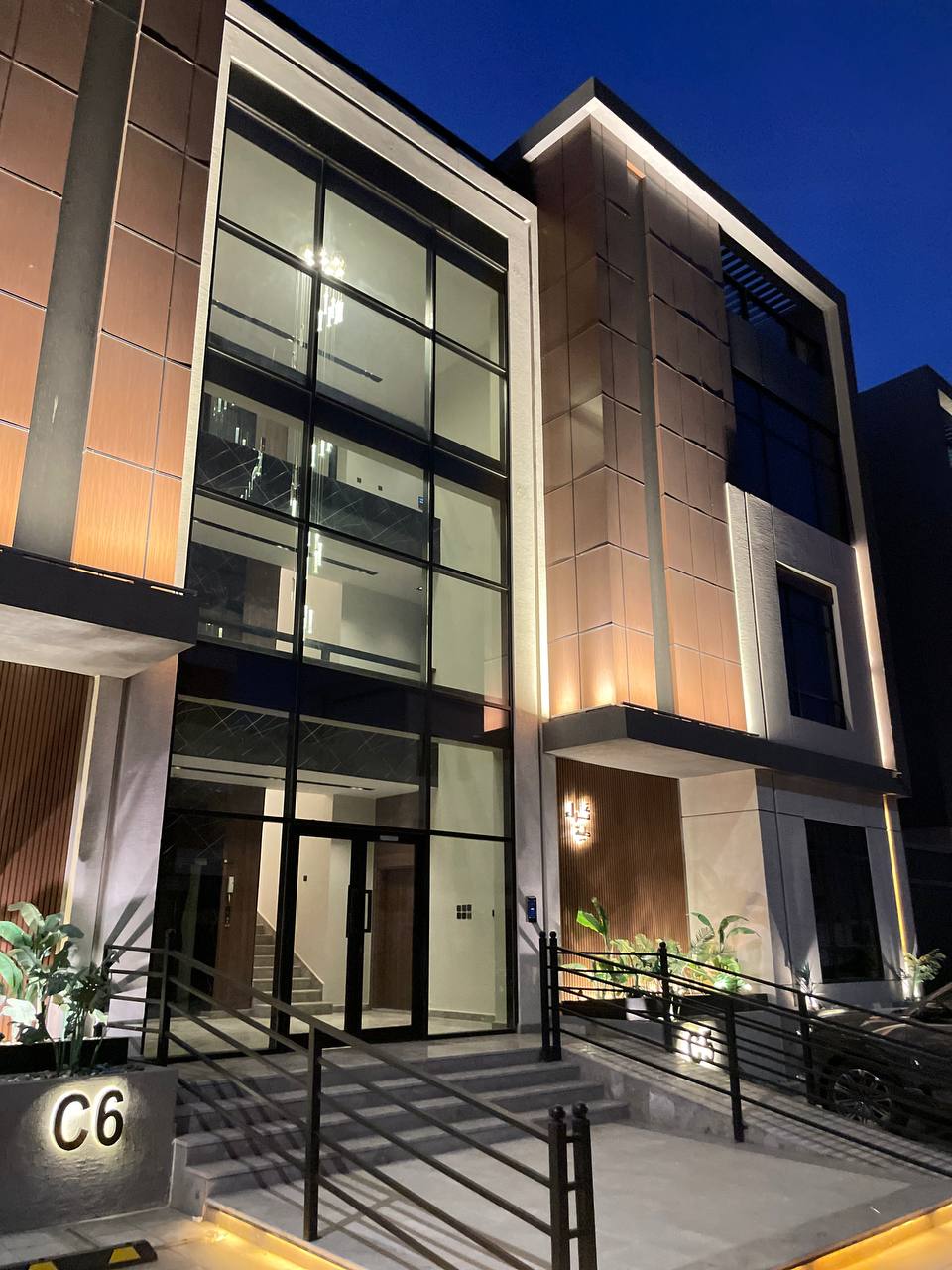 The building exterior features a modern design with large glass windows and warm wooden accents. Soft lighting highlights the architectural details, while green plants are placed at the entrance. A set of steps leads to the main door, providing easy access.