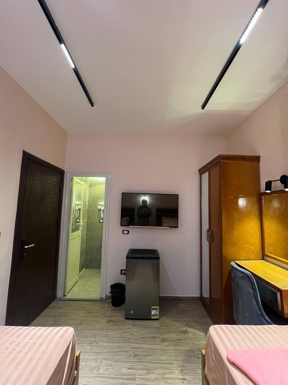 The room features two beds with light pink linens, a wooden wardrobe, and a desk area with a chair. A smart TV is mounted on the wall across from the beds. A private bathroom is accessible through a door, and a mini fridge is positioned beside the desk.