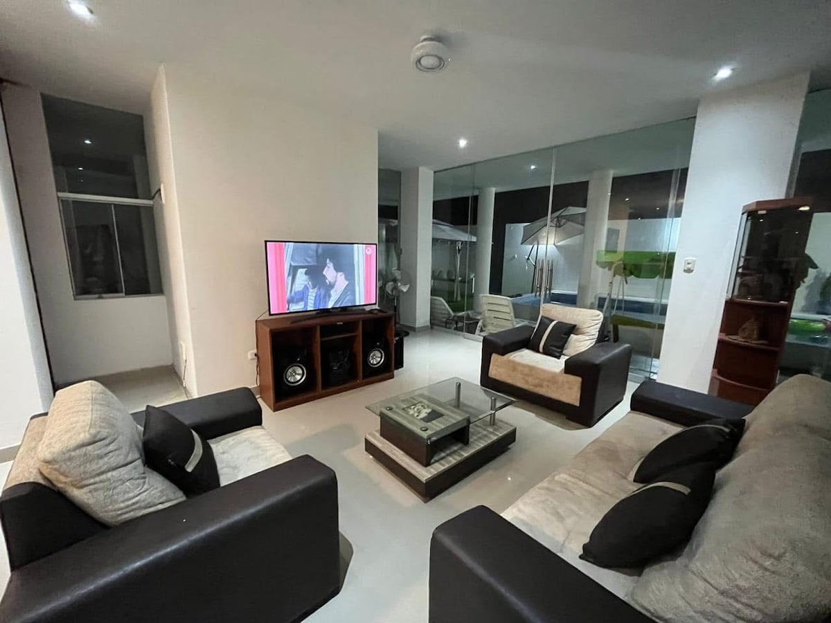A modern living area is featured, furnished with two contrasting sofas, one dark and one light, arranged around a central coffee table. A large television is mounted on a cabinet, while glass walls provide views of the outdoor space.