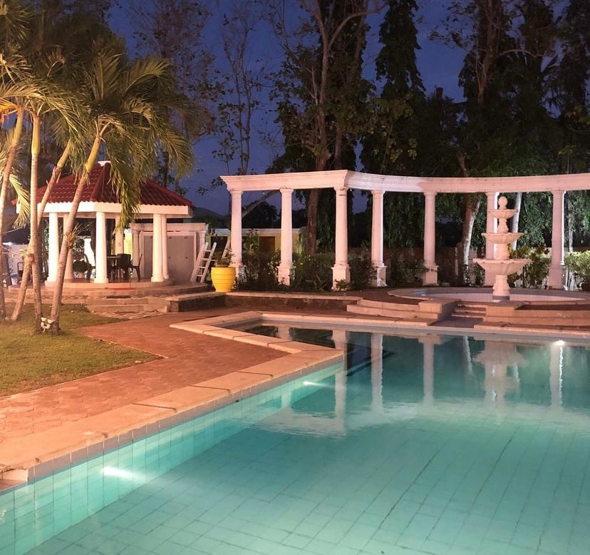 A tranquil pool area is enhanced by a serene fountain and surrounded by well-maintained greenery. A gazebo and seating can be seen nearby, creating inviting spaces for relaxation. Soft lighting illuminates the pool, showcasing its refreshing blue water against the night sky.
