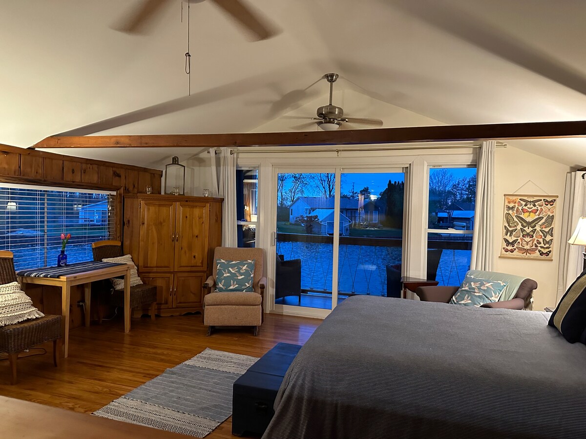 A cozy sleeping area features a spacious bed with light bedding. A wooden table and two chairs are placed beside a large window overlooking the water. Soft lighting enhances the room's warmth, complemented by wooden accents and a fan overhead.