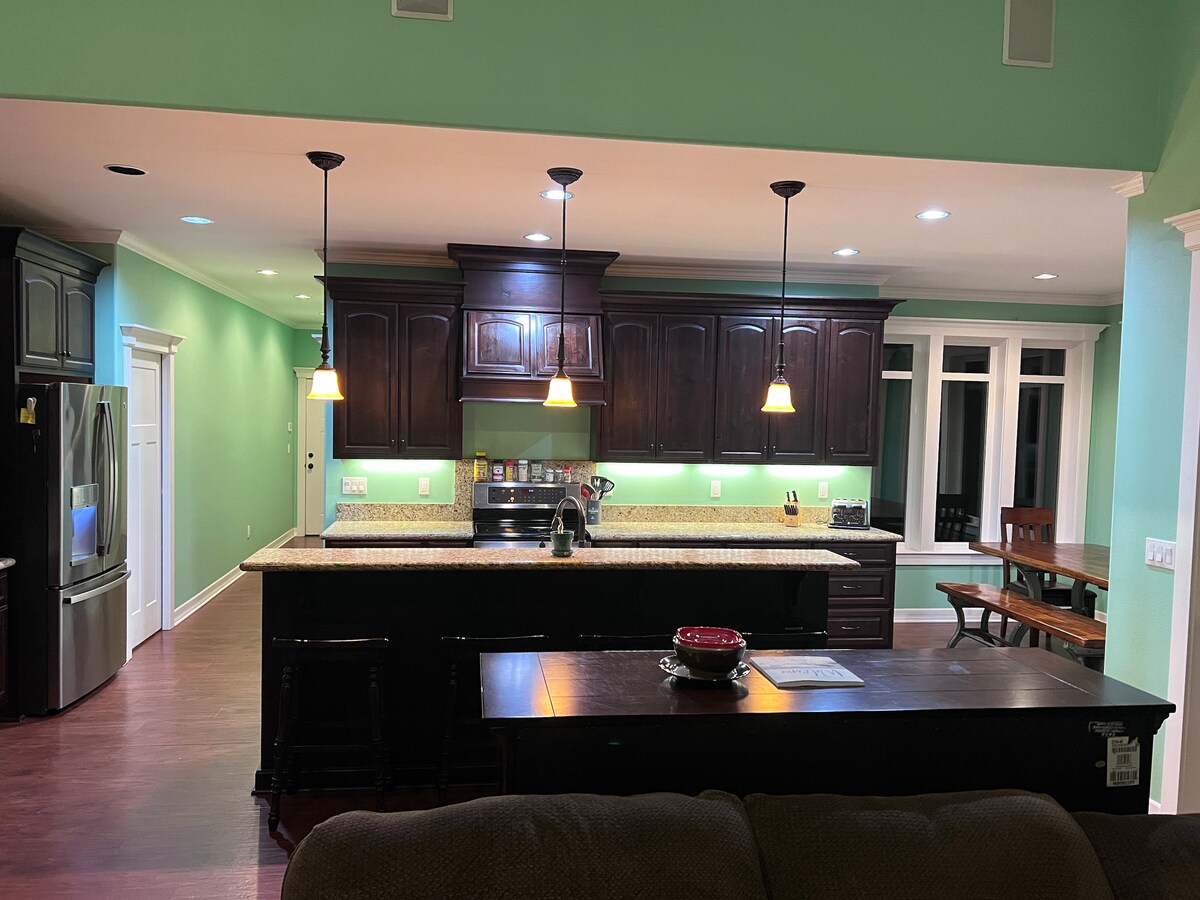 The kitchen features dark wooden cabinets and a spacious granite countertop with pendant lighting overhead. An island bar is positioned centrally, while a large dining table with rustic benches is visible in the background. Appliances such as a refrigerator are present, enhancing functionality.