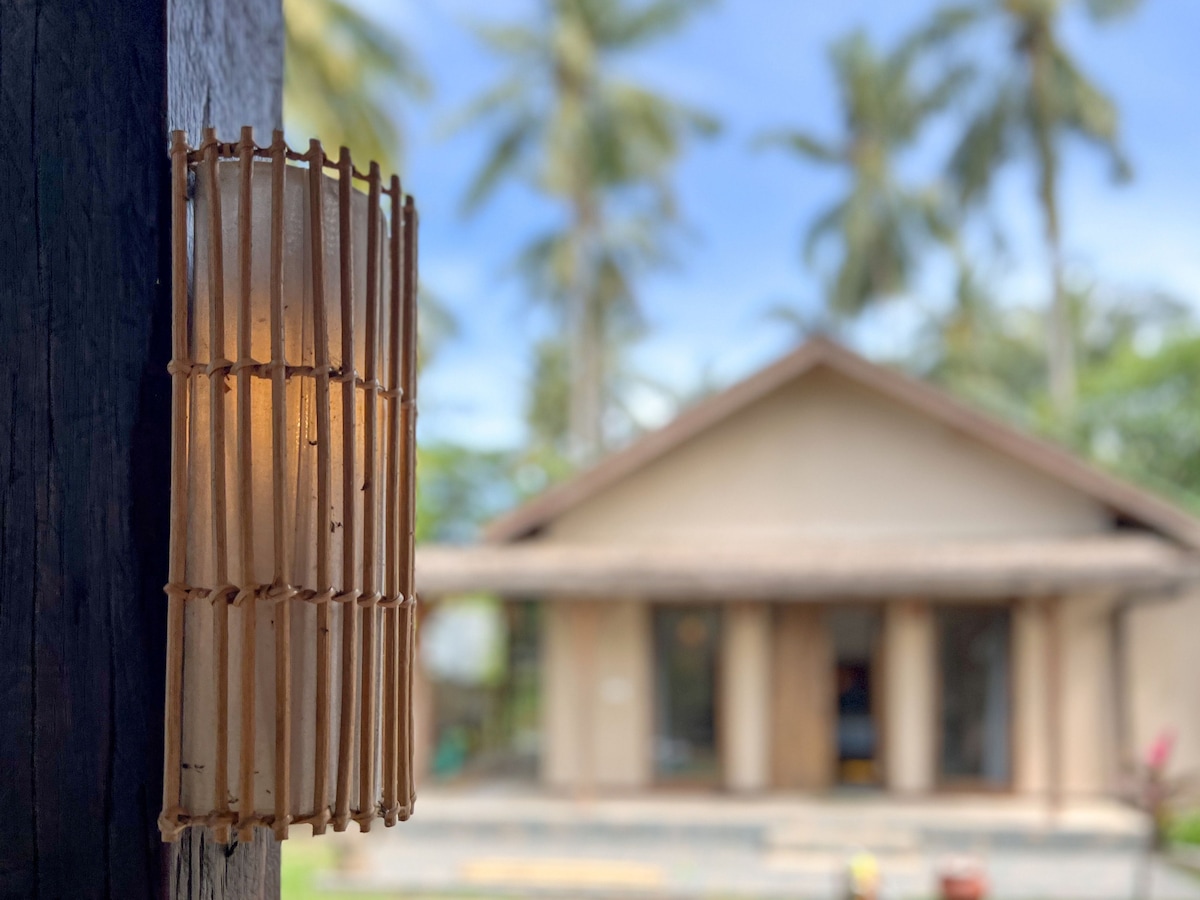 A warm light fixture crafted from natural materials is displayed in the foreground, providing soft illumination. In the background, a serene house is framed by lush palm trees, creating a calming tropical setting.
