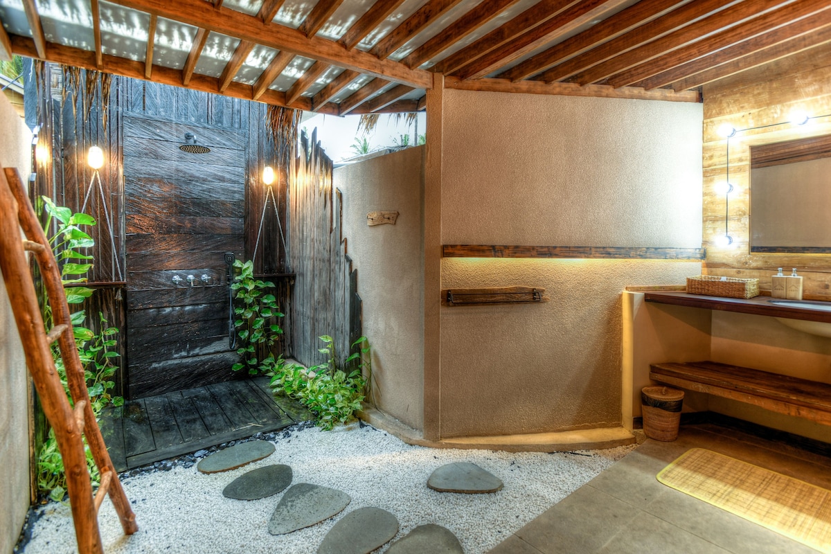 An outdoor shower area is bordered by natural wooden elements and lush greenery. Smooth, flat stones lead through pebble pathways, while warm lighting highlights the rustic decor. A well-organized vanity with a mirror and storage enhances the functional layout of this serene space.