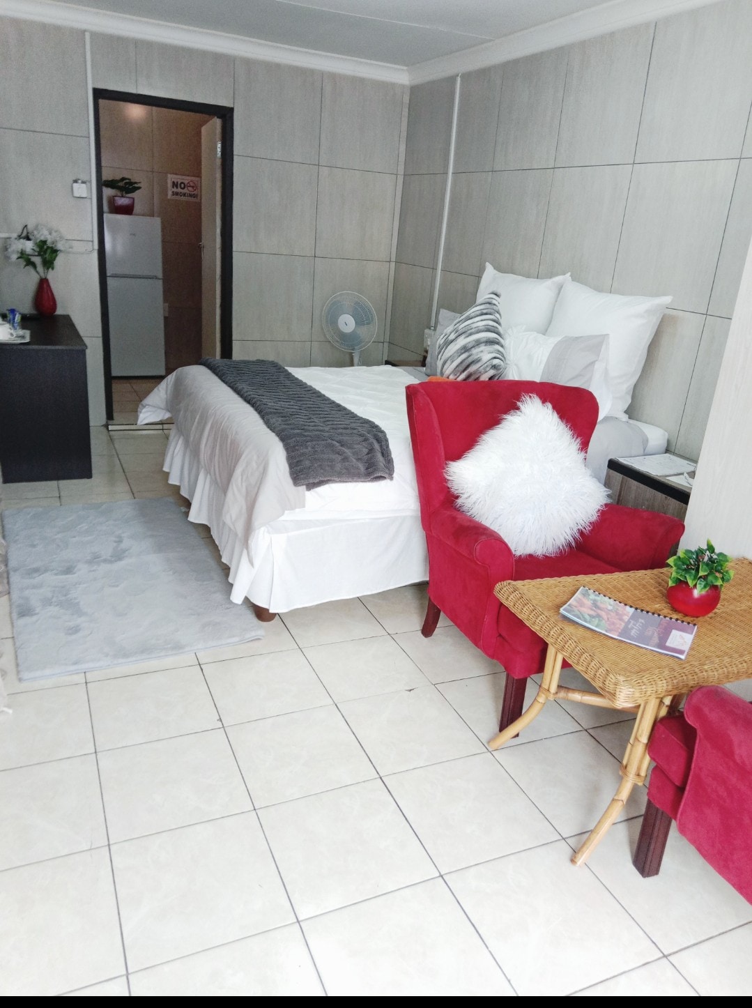 A cozy living space features a spacious bed adorned with neutral-toned linens. Two red armchairs complement a small woven table, while a soft area rug adds warmth. A fan and a small plant provide a touch of comfort, with a refrigerator visible in the background.