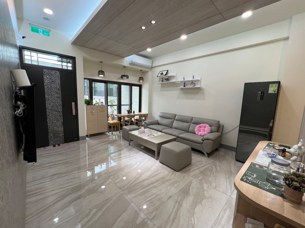 The spacious living area features a modern sofa with a decorative pillow, positioned in front of a minimalist coffee table. Large windows allow for abundant natural light, while an elegant dining table sits in the background. A sleek refrigerator and a side table with decorative items are also visible.