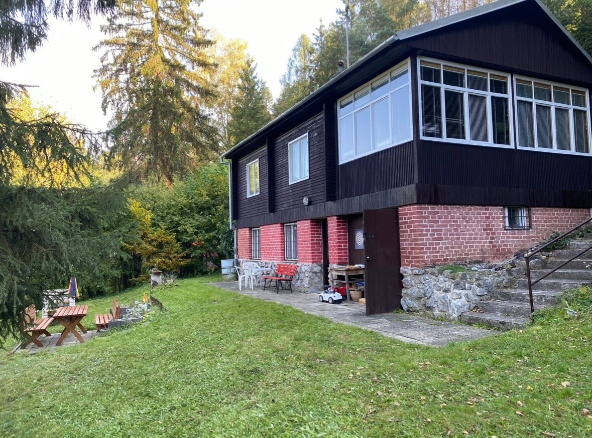 A two-story cabin features a combination of dark wood and red brick siding, surrounded by lush greenery. A patio area with picnic tables and white chairs is positioned on the lawn, leading to a stone pathway. Large windows invite natural light into the living space.