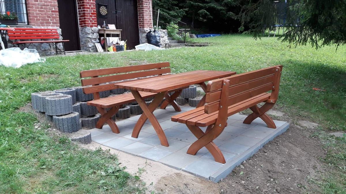 An outdoor dining area features a wooden table surrounded by two matching benches. The seating is situated on a stone-paved surface, with grassy greenery in the background. Wooden accents add warmth to the space, while the arrangement invites relaxation and meals outdoors.