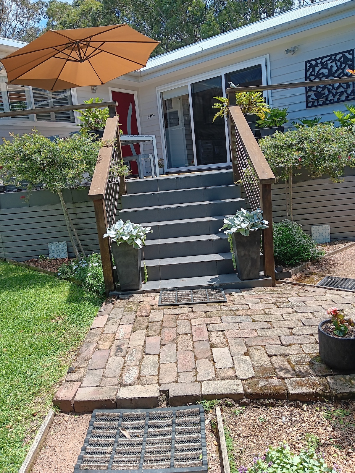 A set of gray steps leads up to the cottage entrance, flanked by planters with greenery. An umbrella provides shade over the stairs, while a pathway of cobblestones connects the area to the surrounding garden, featuring a well-kept lawn and flower beds.