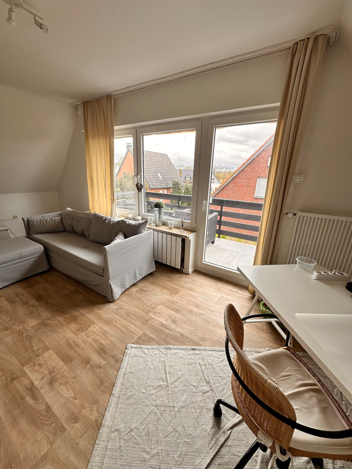 A bright living area features a comfortable sofa and a workspace with a chair. Large glass doors provide access to a balcony, allowing natural light to fill the room. Soft curtains frame the windows, and a neutral rug enhances the cozy atmosphere.