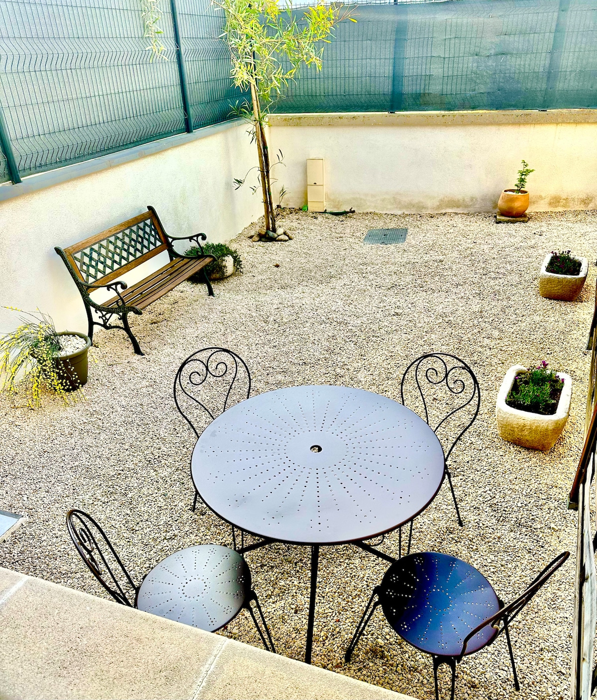 A private outdoor courtyard features a round metal table surrounded by four matching chairs. Planters with small greenery are positioned around the space, and a bench is placed against the wall. A tree adds a natural touch to the gravel-covered area.