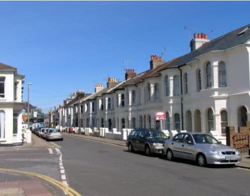 A quiet street is lined with white, two-story houses, featuring brick accents and multiple chimneys. Vehicles are parked along the curb, with a clear blue sky above. The residential area is characterized by its friendly atmosphere and proximity to local amenities.