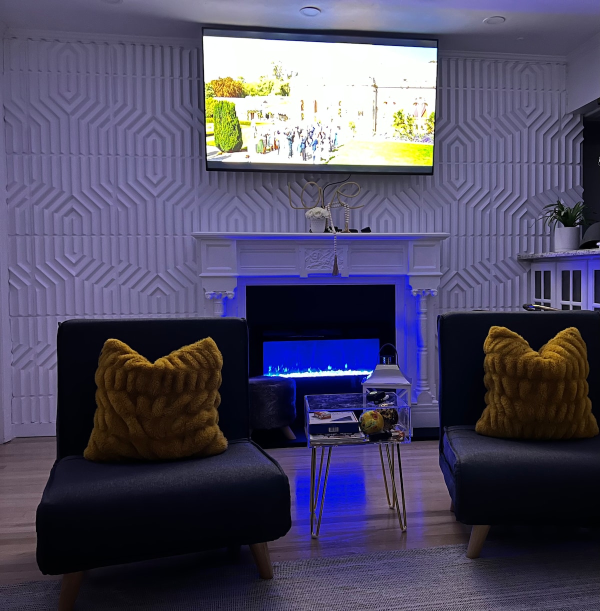 A modern living area features two dark blue chairs with textured mustard cushions, positioned in front of a sleek electric fireplace. A wall-mounted television displays an outdoor scene above a stylish mantel. Soft blue lighting enhances the ambiance of the space.