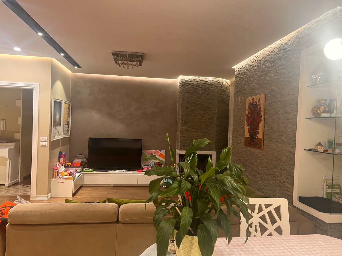 A modern living area features a large flat-screen TV mounted on a white media console. Natural stone accents are seen along the wall, while a green indoor plant adds a touch of nature. A dining table with a geometric design is visible, enhancing the inviting space.