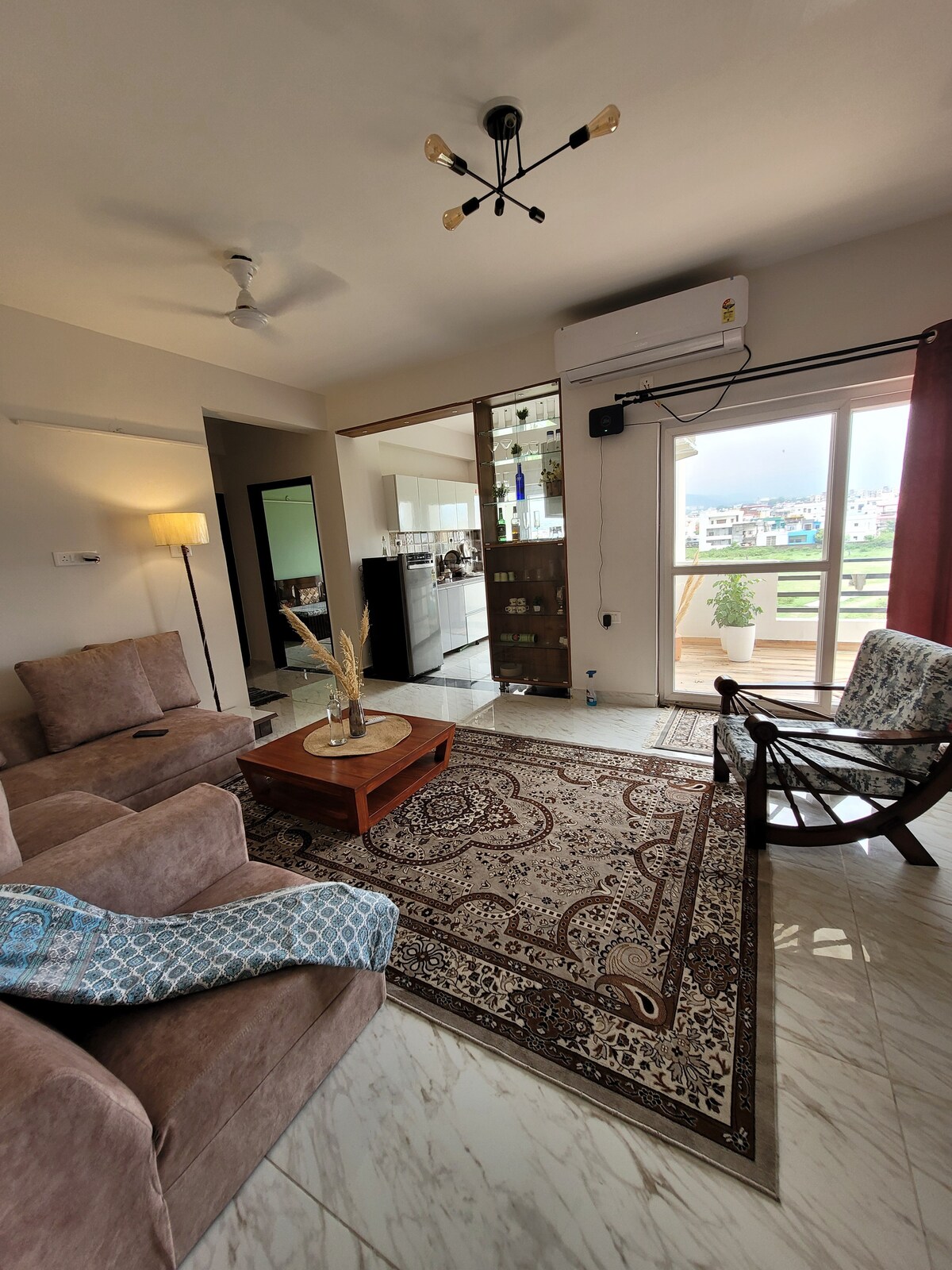 A cozy living area features comfortable seating arrangements, including a plush sofa and an upholstered chair. A wooden coffee table sits on a decorative rug, while large glass doors provide access to the balcony. Natural light enters through the curtains, enhancing the space.