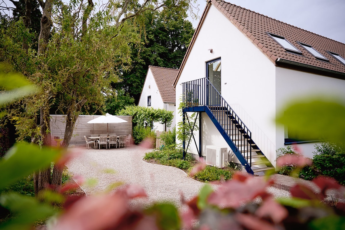 The exterior of a renovated holiday home is visible, featuring a modern white facade with a sloping roof. A blue staircase leads to the entrance, surrounded by greenery. A patio area with a table and chairs is situated nearby, providing a comfortable outdoor setting.