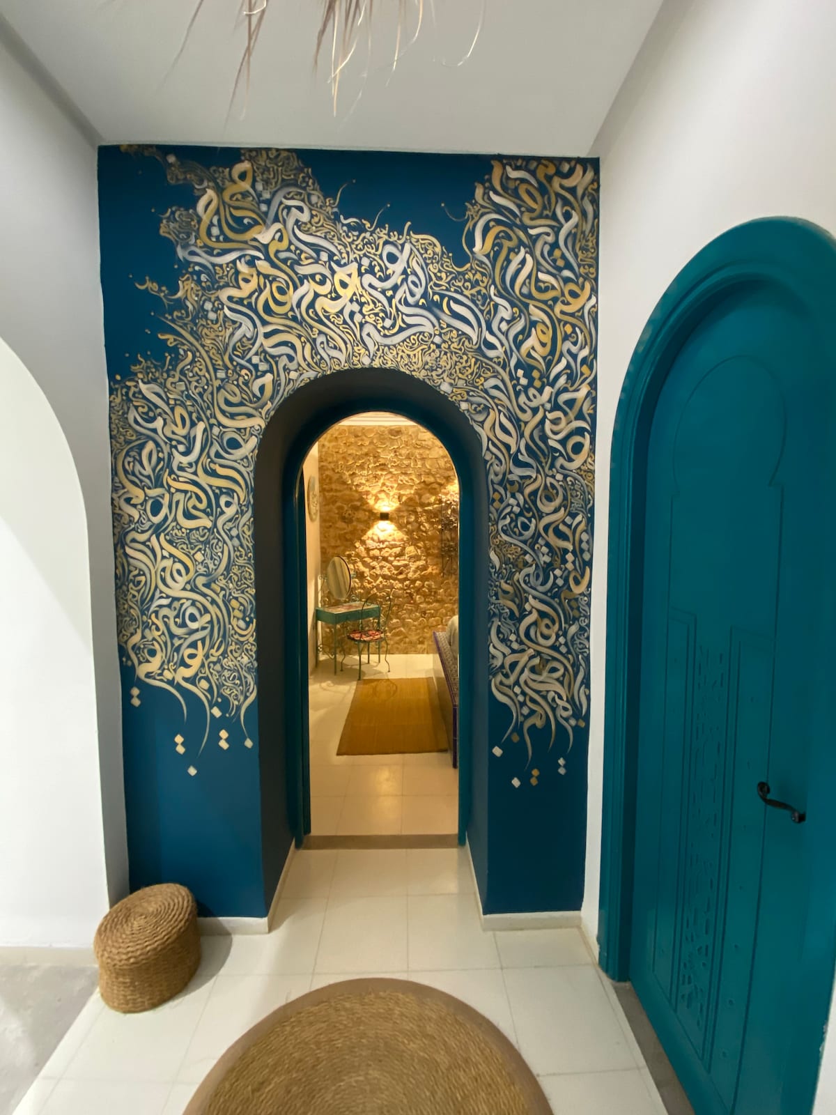 An elegantly decorated hallway features a vibrant blue wall adorned with intricate gold designs. Arched doorways lead to a cozy living space, while a natural fiber stool sits at the base, enhancing the welcoming atmosphere. A soft glow is visible in the distance, adding warmth.