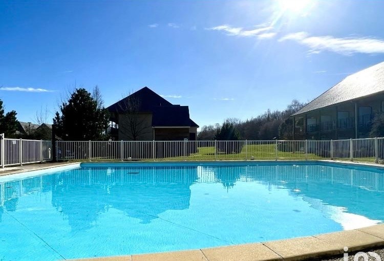 A large outdoor swimming pool is surrounded by a white safety fence, offering a clear view of the bright blue water. Sunlight reflects off the surface, creating a serene atmosphere. In the background, a grassy area and several residential structures are visible.