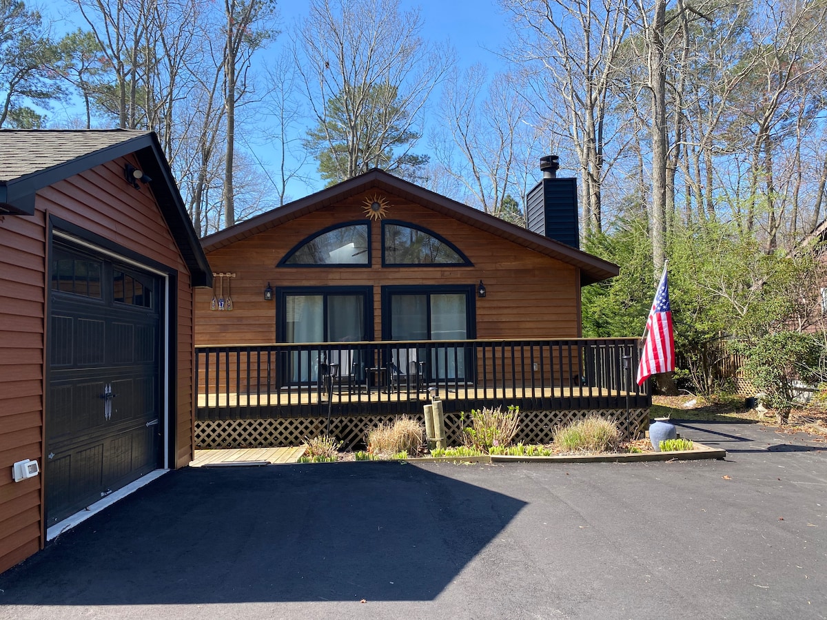 A single-family house features a wooden exterior with dark trim and a large front deck. An American flag is displayed near the entrance, while surrounding trees provide a natural backdrop. The garage, visible on the left, complements the home’s design.