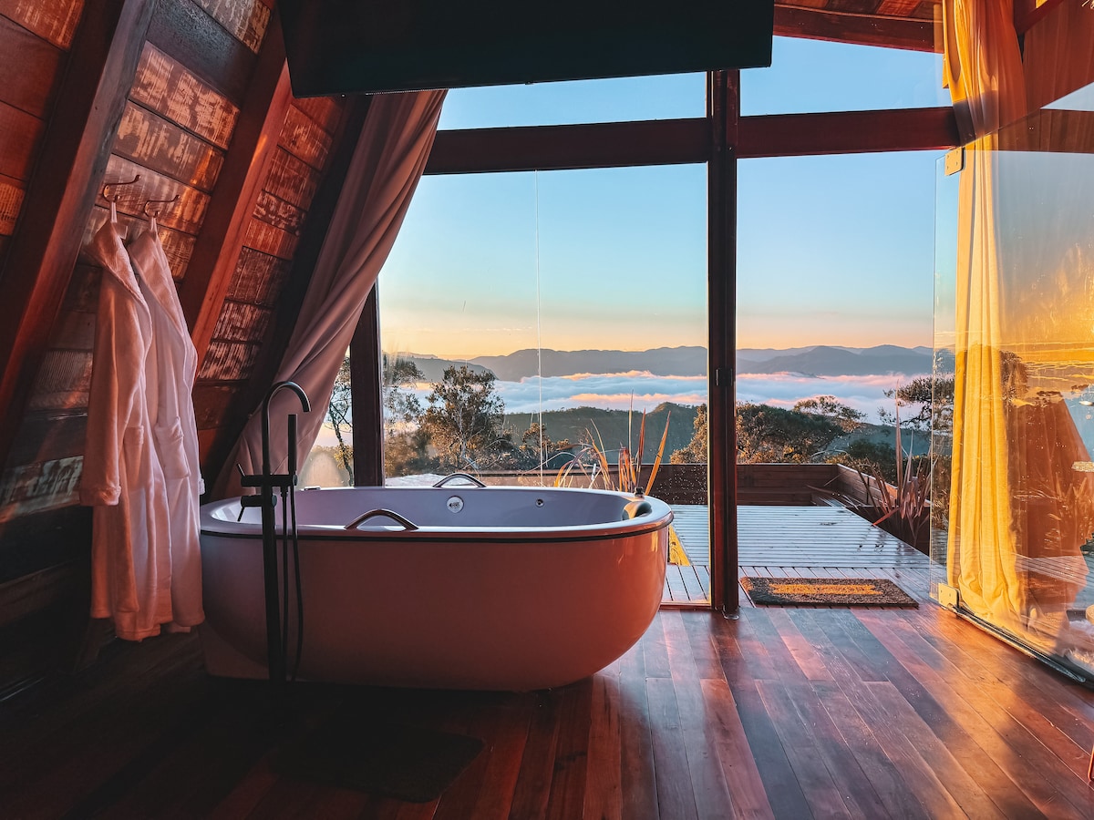 A freestanding bathtub is positioned near large glass windows, offering an unobstructed view of the mountain landscape at sunrise. Natural light fills the space, while warm wood tones create a cozy ambience. A plush robe hangs nearby, enhancing the sense of comfort.