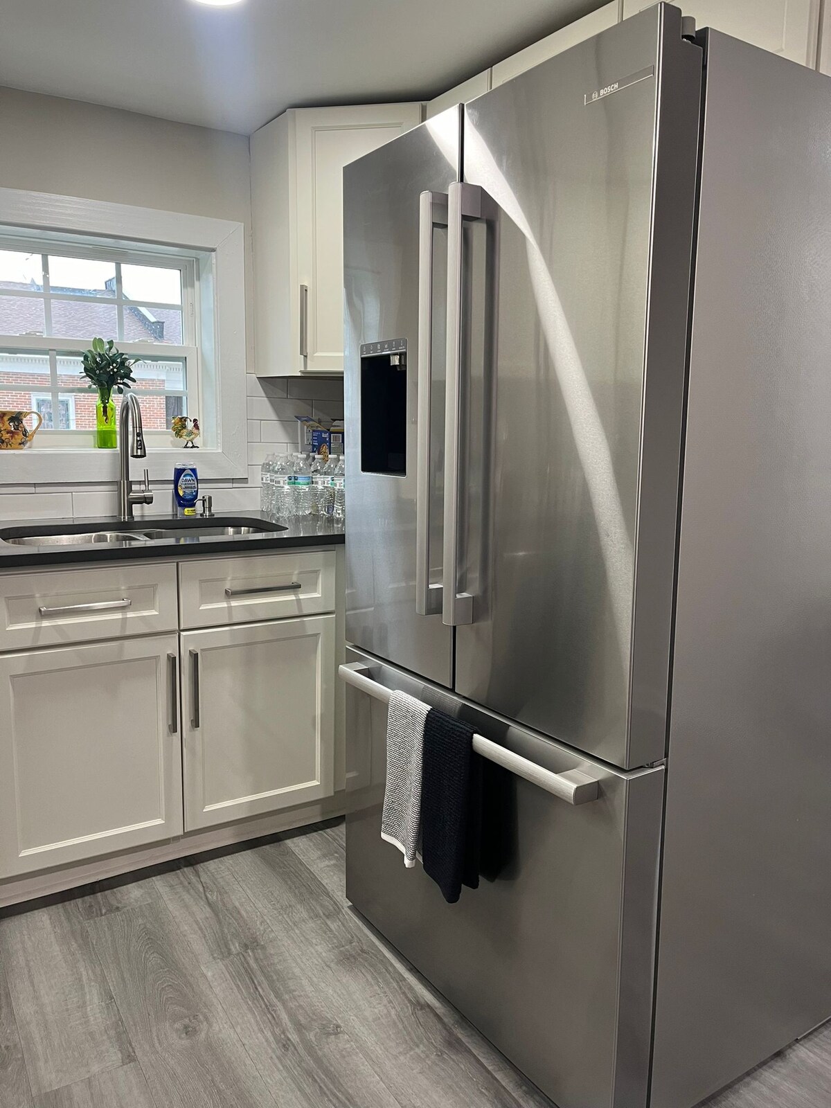A modern kitchen showcases stainless steel appliances, including a large refrigerator with a convenient water dispenser. White cabinetry complements the sleek counters. A window offers natural light, while a small plant adds a touch of greenery to the space.