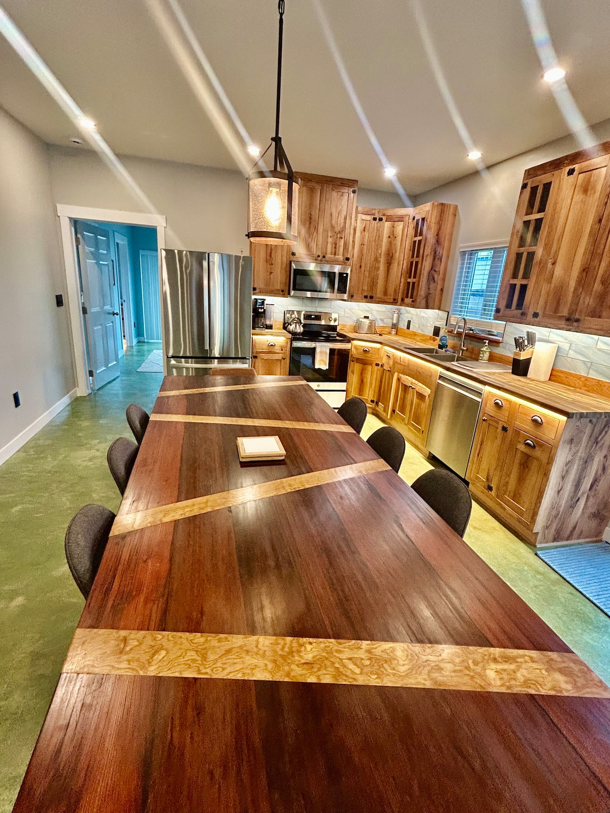 A spacious kitchen area features custom wooden cabinetry and modern appliances. A large handmade dining table with unique inlay sits in the center, surrounded by comfortable chairs. Soft lighting highlights the rich wood tones, while natural light streams in through the window.