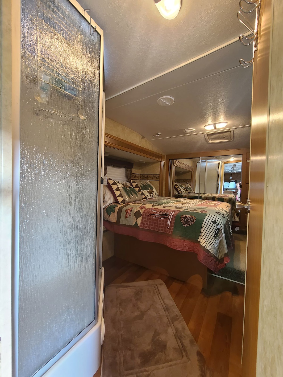 A narrow hallway leads to a cozy sleeping area featuring a queen bed dressed in a patterned quilt. The space includes mirrored cabinetry, providing a sense of openness. Light illuminates the hallway, highlighting rich wood flooring and soft carpeting.