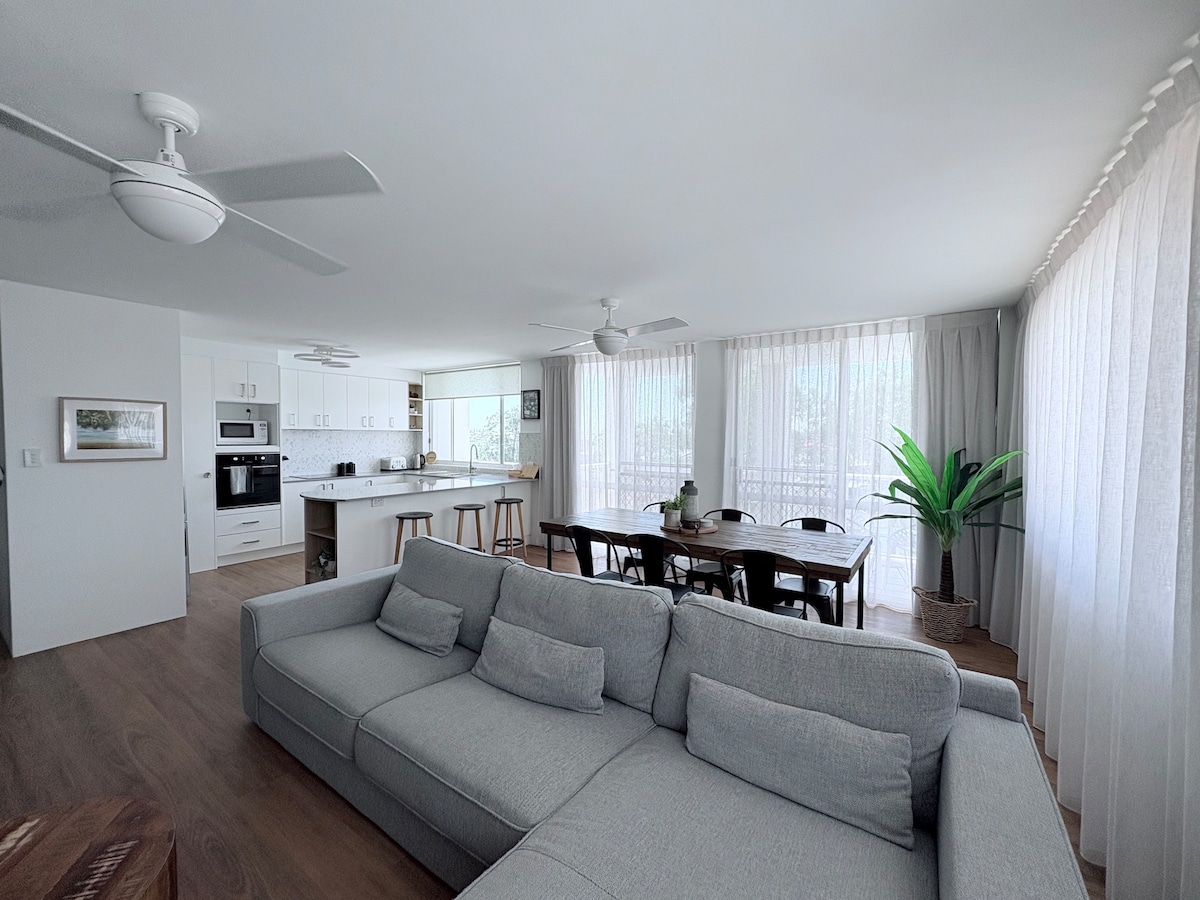 An open-plan living area features a comfortable grey sectional sofa, complemented by a wooden dining table. Bright natural light filters through sheer curtains, illuminating the space. A large plant adds a touch of greenery, while modern kitchen appliances are visible in the background.