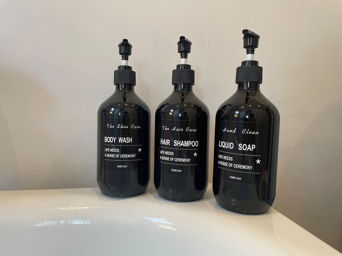 Three matching black dispensers are arranged on a bathroom counter. Labels indicate 'Body Wash,' 'Hair Shampoo,' and 'Liquid Soap.' Each dispenser features a pump top, providing a modern and coordinated look for bathroom essentials.