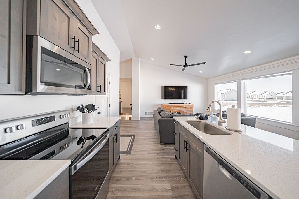 A modern kitchen features sleek cabinetry, a large island with a sink, and black appliances, including a microwave and stovetop. Natural light floods the space from spacious windows overlooking the living area, where a gray sofa and a mounted TV create a welcoming ambiance.