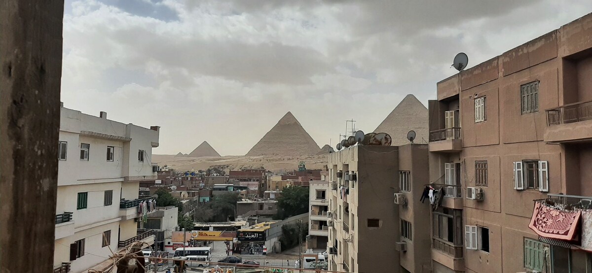 A view of the Pyramids of Giza is captured from a balcony, framed by neighboring apartment buildings. The scene presents a mix of structures alongside a bustling street, with laundry hanging in the distance. The pyramids rise prominently against a cloudy sky.