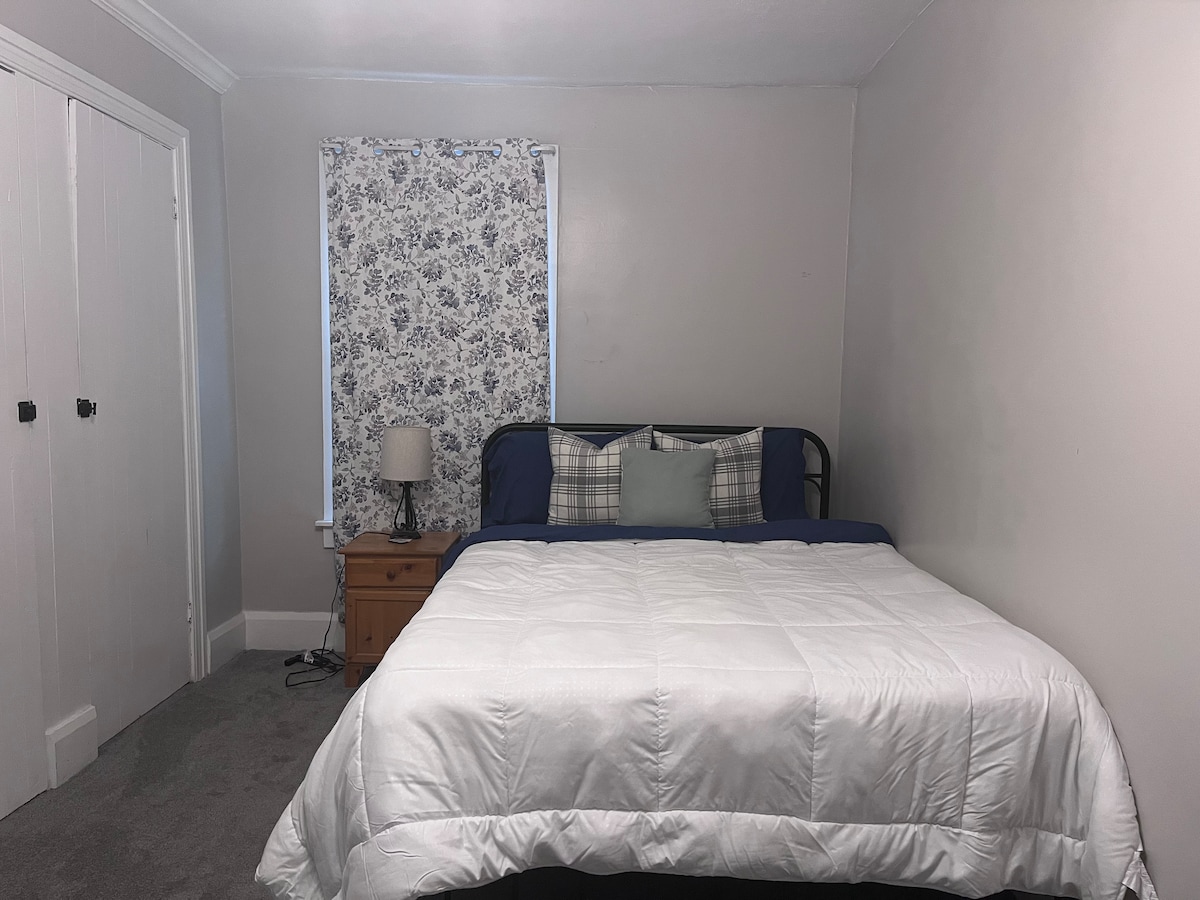 A private bedroom features a spacious bed dressed in a white quilt. Pillows in various patterns rest against a backdrop of a floral-patterned window treatment. A small bedside lamp sits atop a bedside table next to the bed, with a light gray wall and carpeted floor completing the room.
