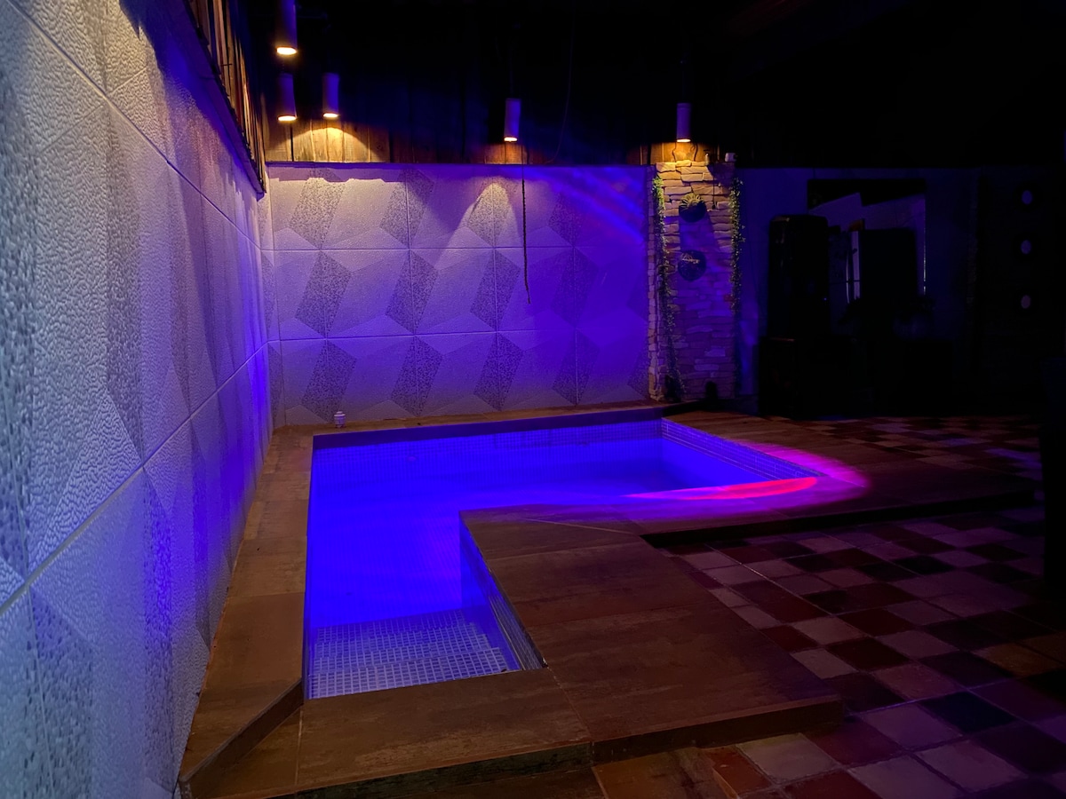 An inviting pool area is illuminated with calming blue and purple lights. The pool features a modern design with steps leading into the water, surrounded by textured walls and stone accents. Pendant lights hang above, enhancing the serene evening atmosphere.