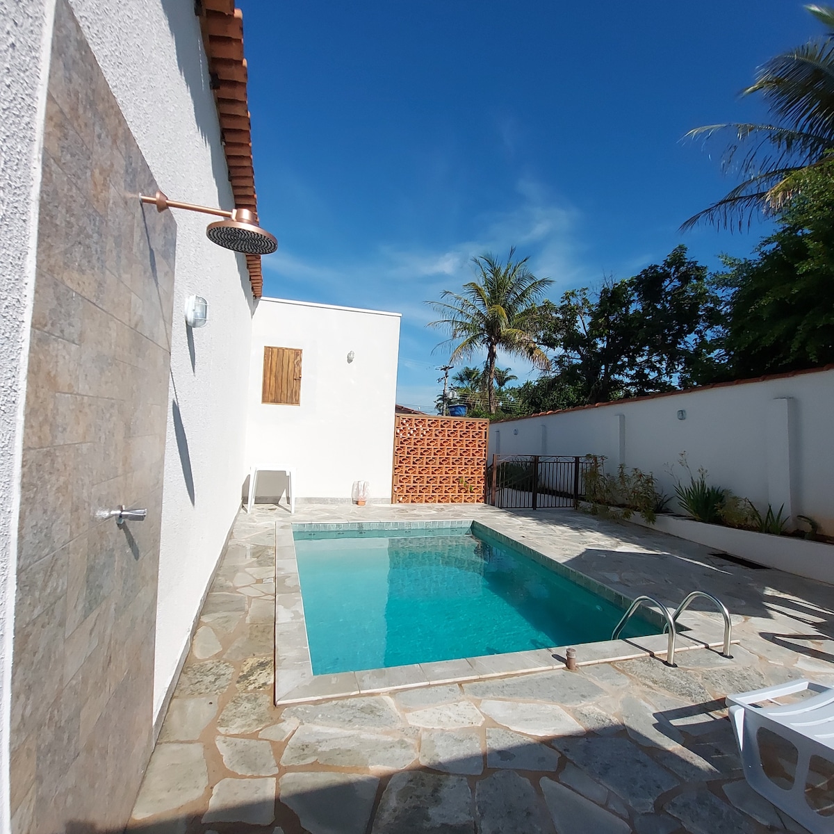 A private outdoor area features a clear swimming pool with a stone border and a surrounding patio. Sunlight reflects on the water, and tropical plants are visible in the background. A shower is mounted on the nearby wall, adding to the pool's convenience.