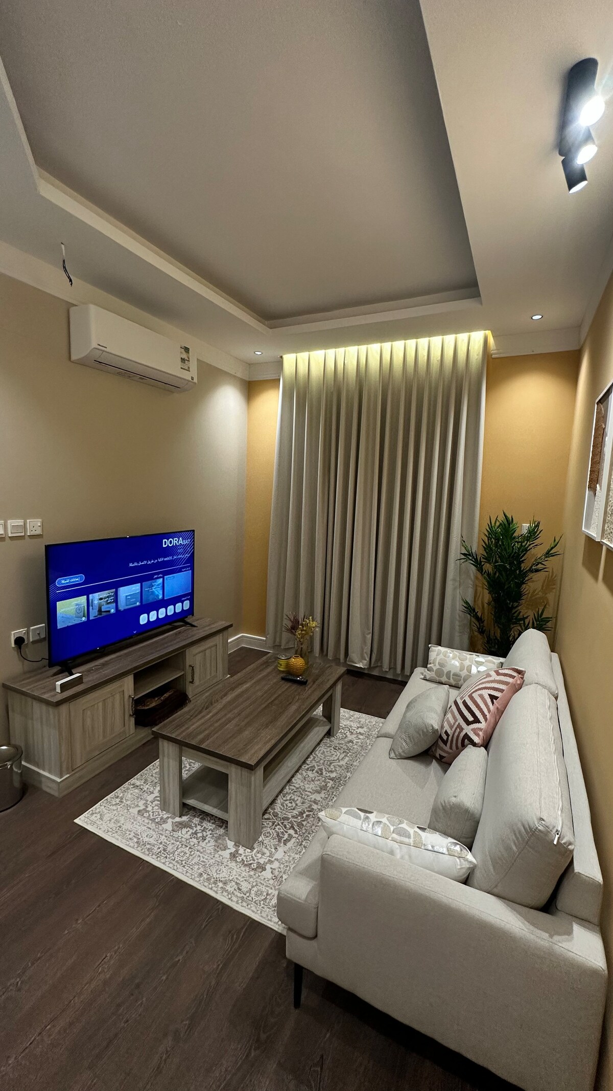A modern living area is highlighted by a comfortable sofa with decorative cushions, positioned next to a wooden coffee table. A flat-screen television is mounted on the wall, and sheer curtains frame a window, allowing natural light to brighten the space.