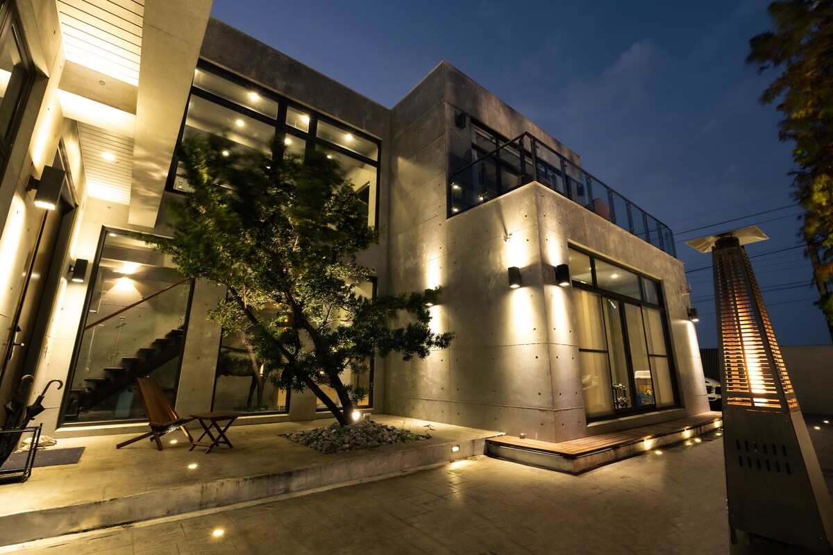 The modern villa exterior is illuminated by soft lighting, showcasing a minimalist design with large glass windows and clean lines. A wooden deck leads to the entryway, and a single tree is placed strategically in the landscaping, enhancing the overall aesthetic.