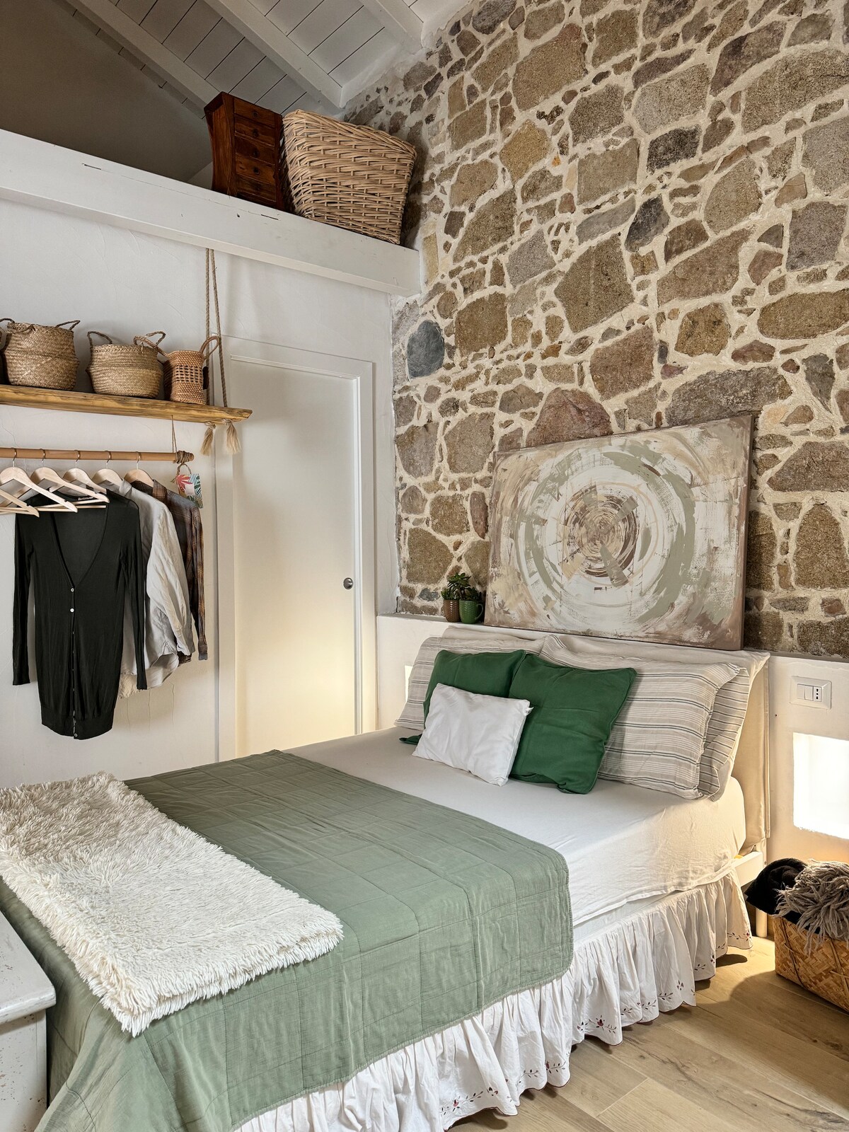 A cozy sleeping area features a spacious bed dressed in white linens with a green throw and accent pillows. Natural stone walls add warmth to the room, complemented by simple decor items on a shelf and a subtle wall art piece above the bed.