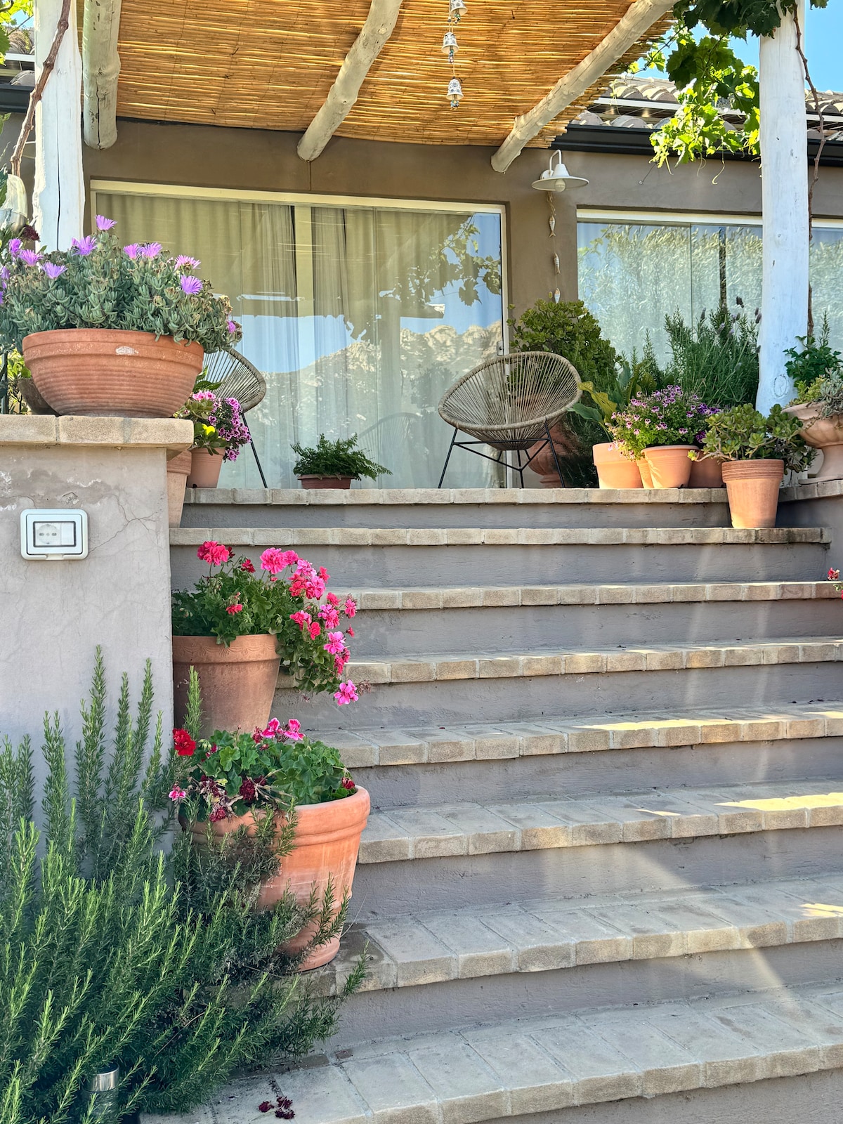 A serene entrance is marked by a set of stone steps leading up to a covered porch. Decorative potted plants, including colorful flowers, frame the pathway, enhancing the inviting atmosphere. Two comfortable chairs are arranged on the porch, providing a cozy spot to enjoy the surroundings.