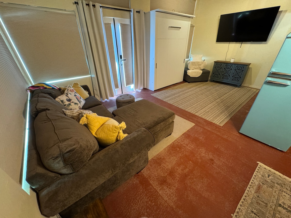 A comfortable seating area is featured, showcasing a sectional couch adorned with bright throw pillows. A wall bed is neatly stowed away, with a large smart TV mounted nearby. Natural light filters in from the glass doors leading outside, complementing the muted color scheme.