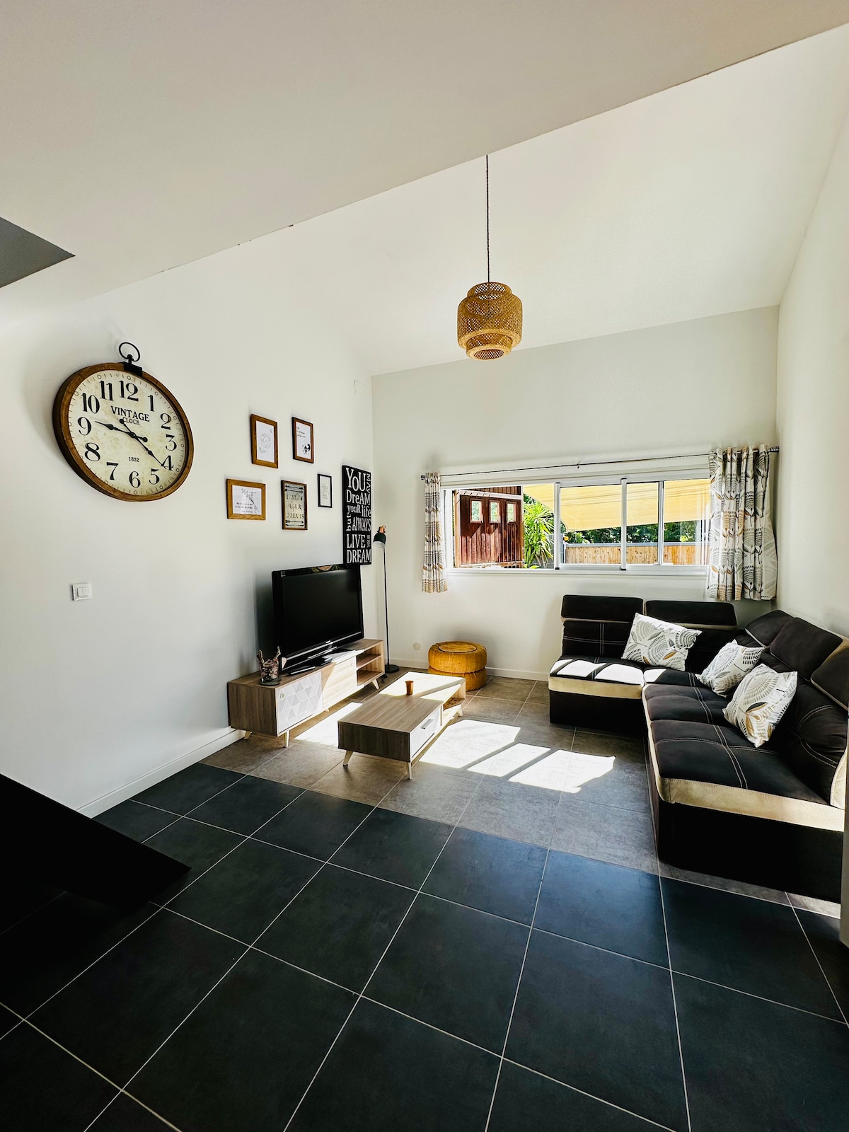 A bright living area is showcased with large windows allowing natural light to fill the space. A cozy seating area features a dark sofa and a wooden coffee table, while a wall clock and framed art add character to the room.