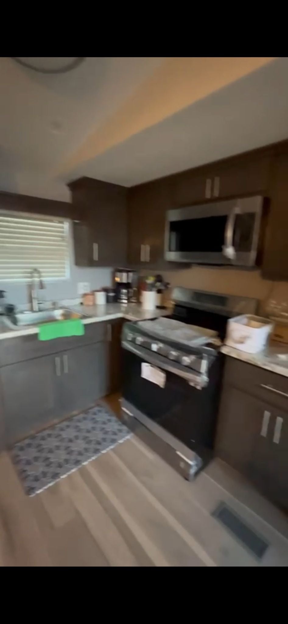 The kitchen features dark cabinetry, modern appliances, and a well-organized counter space. A stove and microwave are prominently displayed, with a sink positioned near a window. A small rug adds comfort to the floor, complementing the overall functionality of the space.