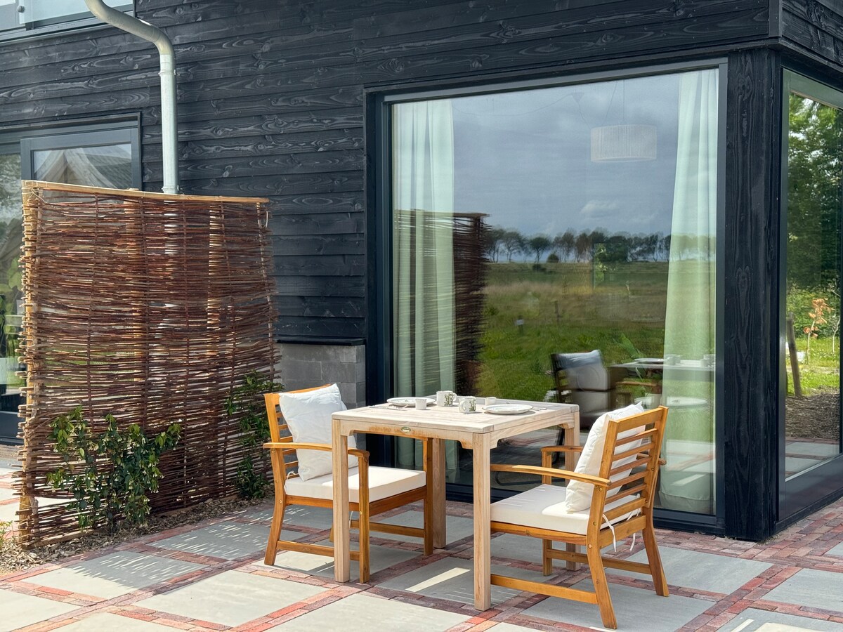 An outdoor dining area features a wooden table with two chairs and a linen table setting. Nearby, a woven privacy screen offers separation while plants add greenery, enhancing the connection to the spacious natural surroundings visible through expansive glass doors.