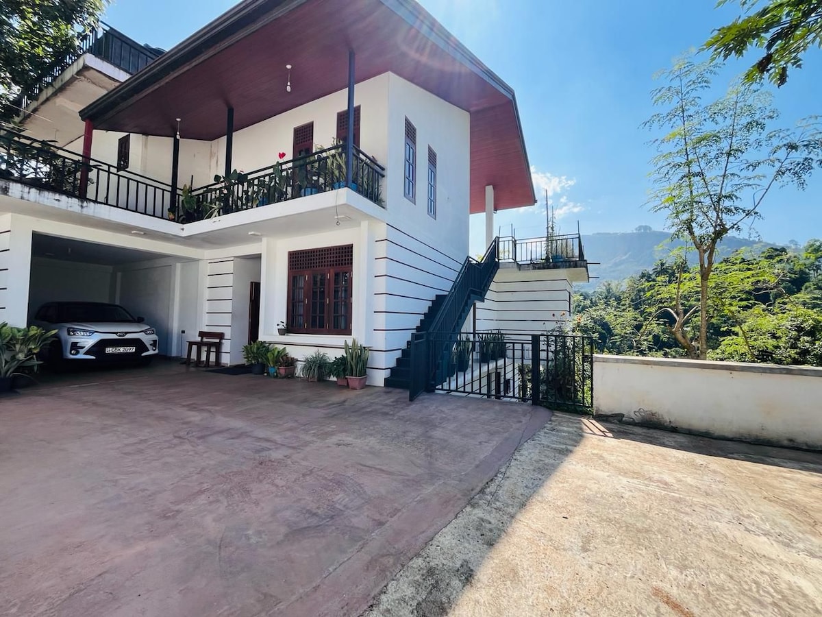 The exterior of a two-story house is presented, showcasing a spacious parking area with a vehicle. A staircase leads to the upper level, while a balcony offers views of the surrounding greenery and mountains. Sunlight bathes the architectural details.