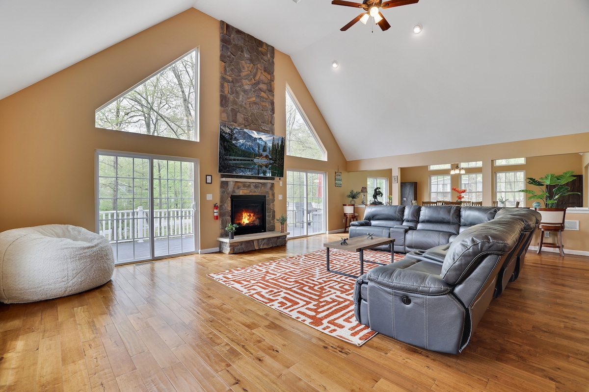 A spacious living area features large windows allowing natural light, with views of surrounding greenery. A stone fireplace serves as a focal point, complemented by a comfortable sectional sofa and a cozy bean bag. Wooden floors add warmth to the modern design.