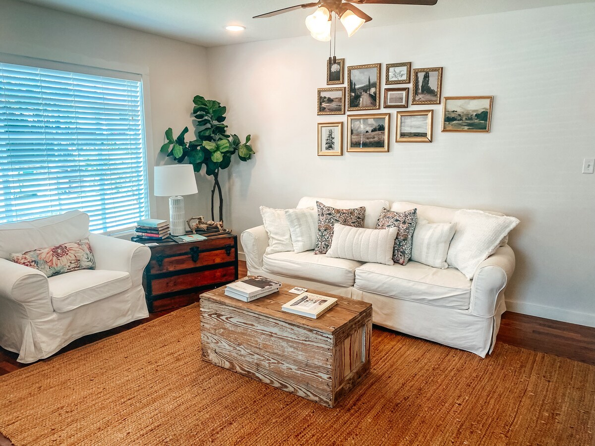 A cozy living room features a white sofa adorned with patterned pillows, accompanied by a white armchair. A rustic wooden coffee table sits at the center, displaying books and decor. Natural light streams through a window covered with sheer blinds, enhancing the welcoming atmosphere.