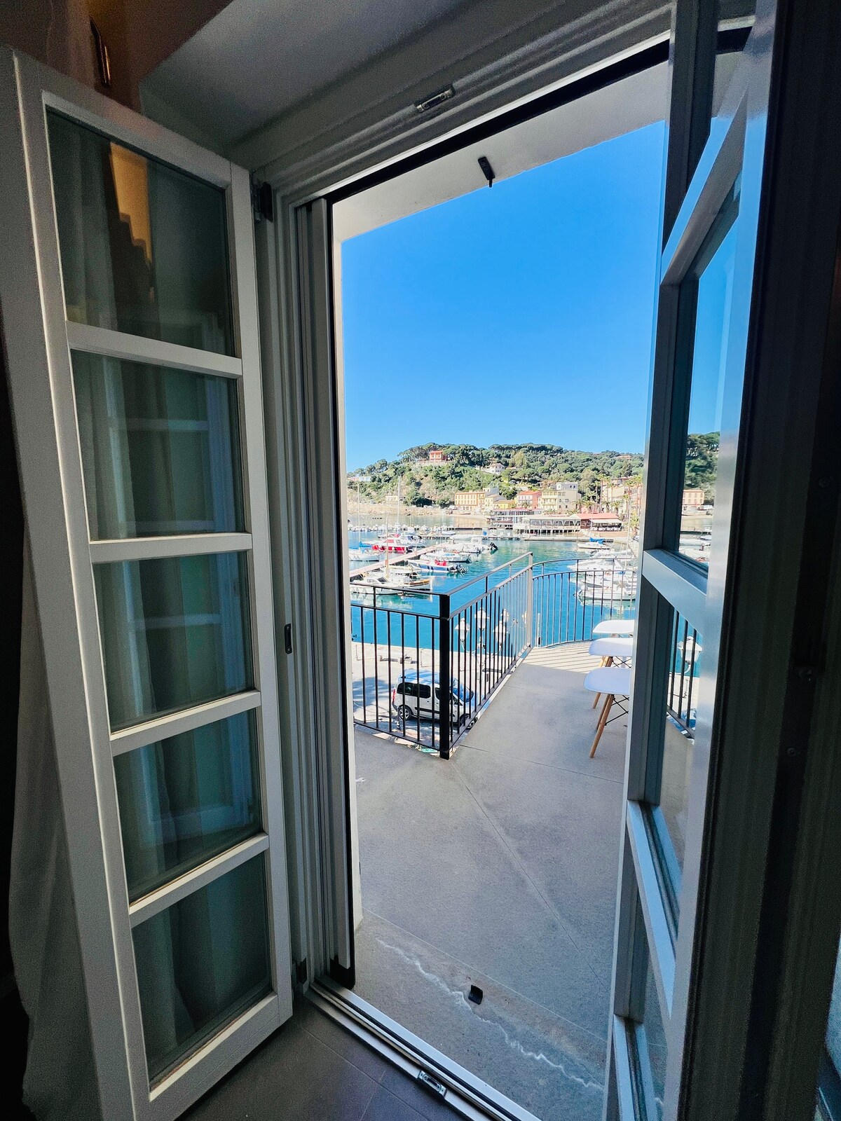 A wide-open door reveals a scenic view of the harbor, with boats gently swaying in the water. A small balcony is visible, featuring a table and chair set against a backdrop of colorful buildings and lush greenery.